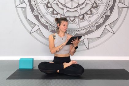 Smiling woman in lotus pose on yoga mat holding a tablet, representing online yoga practice at home.