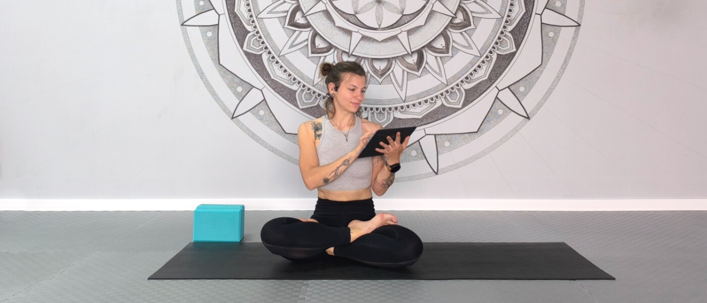 Smiling woman in lotus pose on yoga mat holding a tablet, representing online yoga practice at home.
