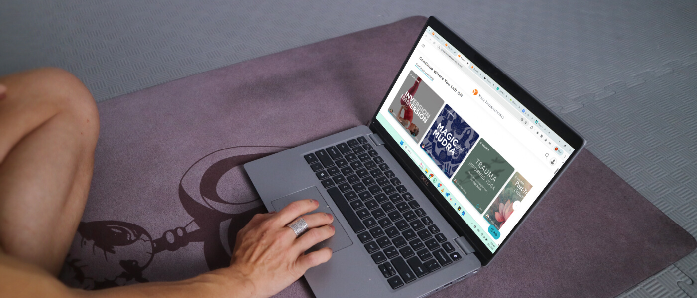 Yoga international website open in browser on a laptop, set on a yoga mat, with a hand selecting the course.