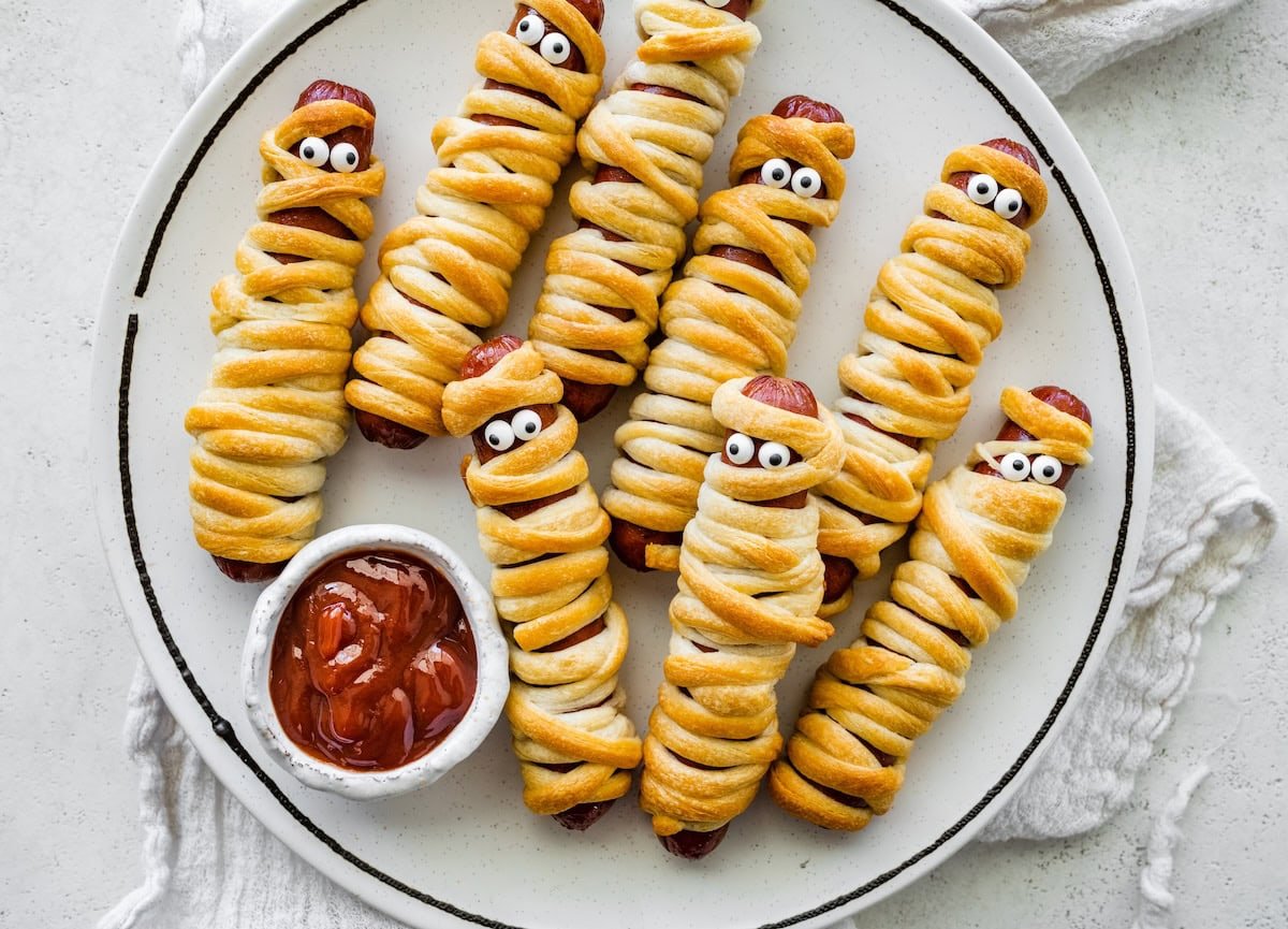 Plate of baked mummy hot dogs wrapped in crescent roll dough with candy eyes, served with ketchup for dipping.