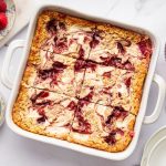 Baked oatmeal in a white baking dish showing creamy cottage cheese and raspberry jam swirls on top. Fresh raspberries and rolled oats are next to the dish.