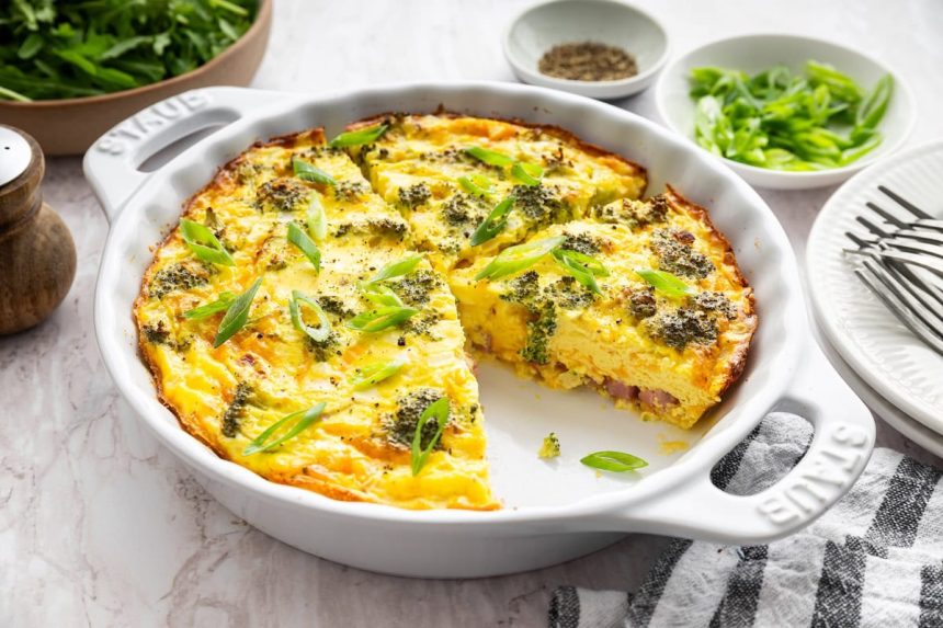 Crustless quiche with broccoli, cheese, onion, and a green onion garnish in a round baking dish with a slice removed.