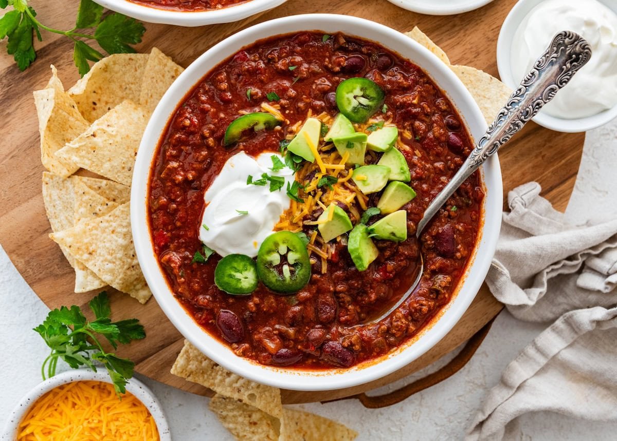 A bowl of slow cooked bean and beef chili topped with avocado, cheese, sour cream, and jalapeno slices.